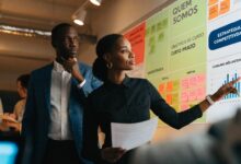 Profissional em ambiente de trabalho colaborativo analisando metas e resultados em um quadro, com colegas ao fundo, representando estratégias para lidar com a competição e crescer na carreira.