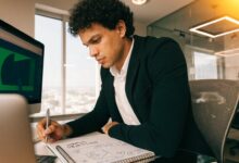 Profissional brasileiro organizando metas de carreira em um caderno diante de um computador, em um ambiente de trabalho iluminado e moderno.
