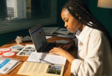 Estudante universitária analisando opções de cursos e instituições em um laptop, com caderno de anotações e materiais acadêmicos sobre a mesa.