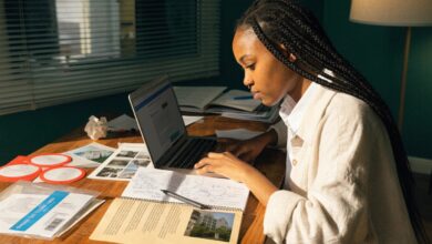 Estudante universitária analisando opções de cursos e instituições em um laptop, com caderno de anotações e materiais acadêmicos sobre a mesa.